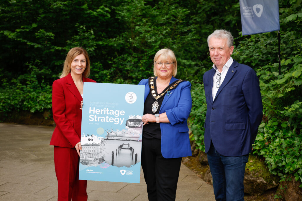Image of three figure (Interim Chief Executive of ABC Council, Lord Mayor of ABC Borough and Dr. Paul Mullan from Heritage Fund) in front of a green background holding a large cover of the Heritage Strategy